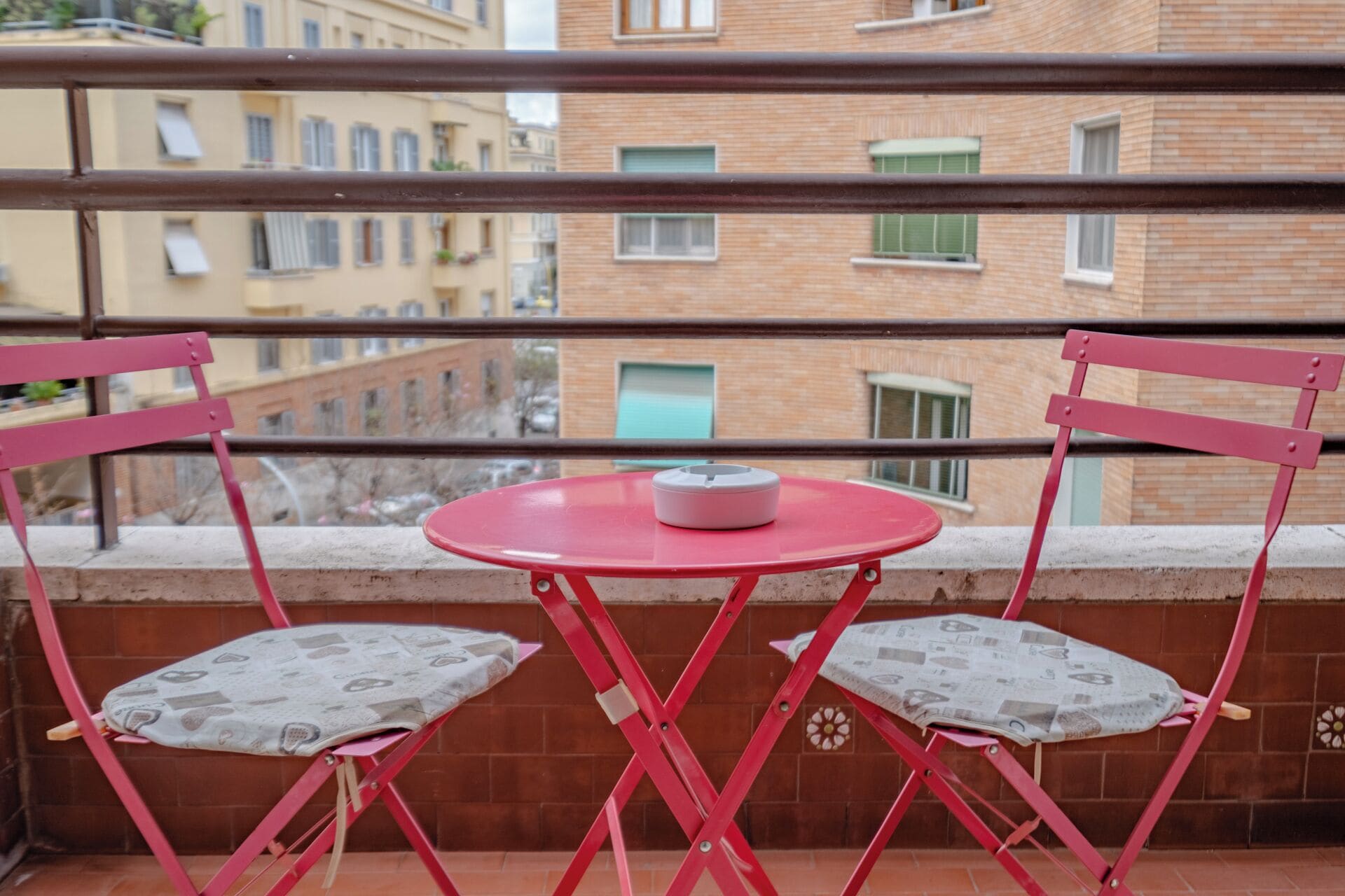 A balcony with two red folding chairs and a small round table, overlooking a cityscape with brick buildings and windows.