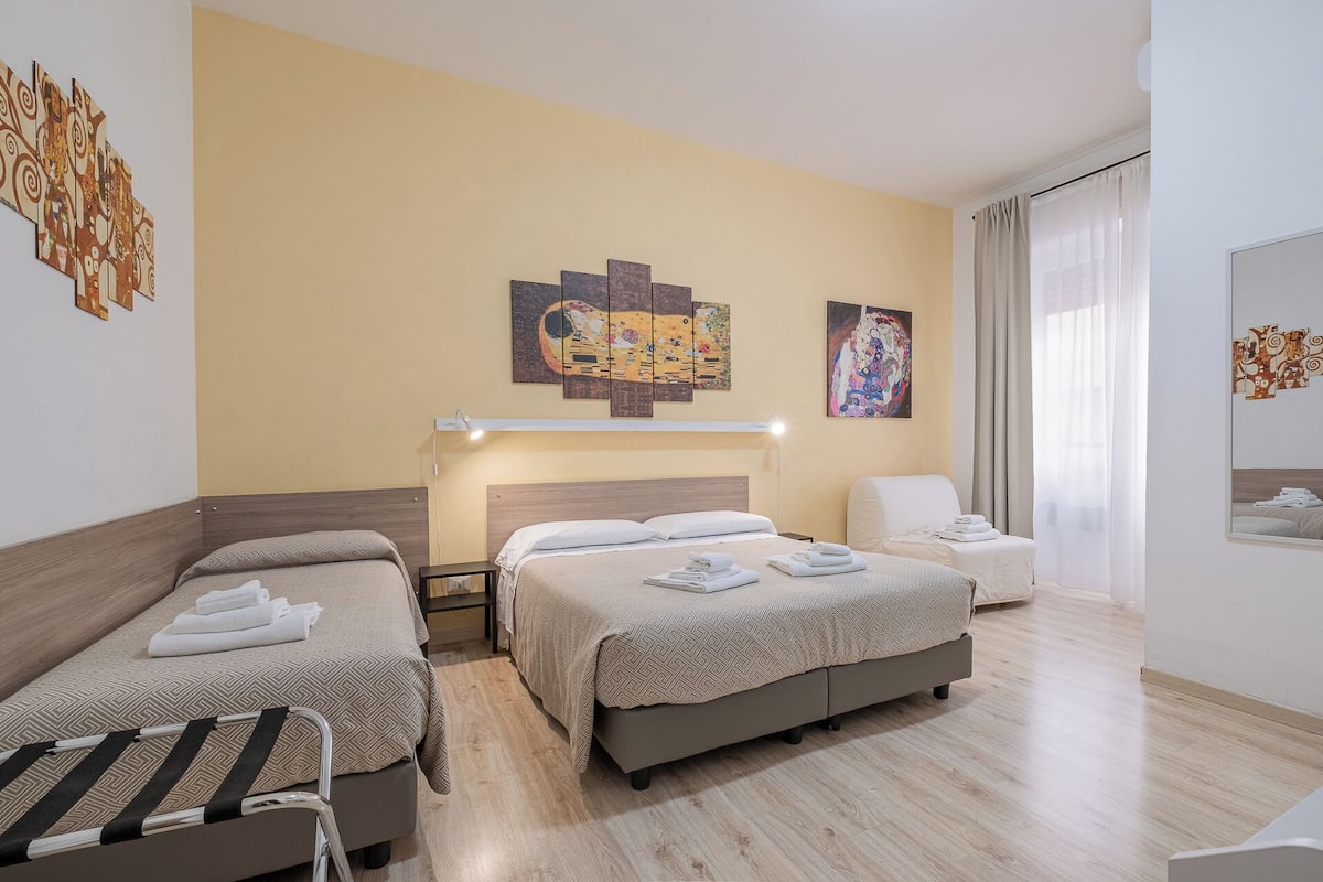 Ein Hotelzimmer mit zwei Betten, einem Nachttisch, einem Sessel und Kunstwerken an den Wänden.
