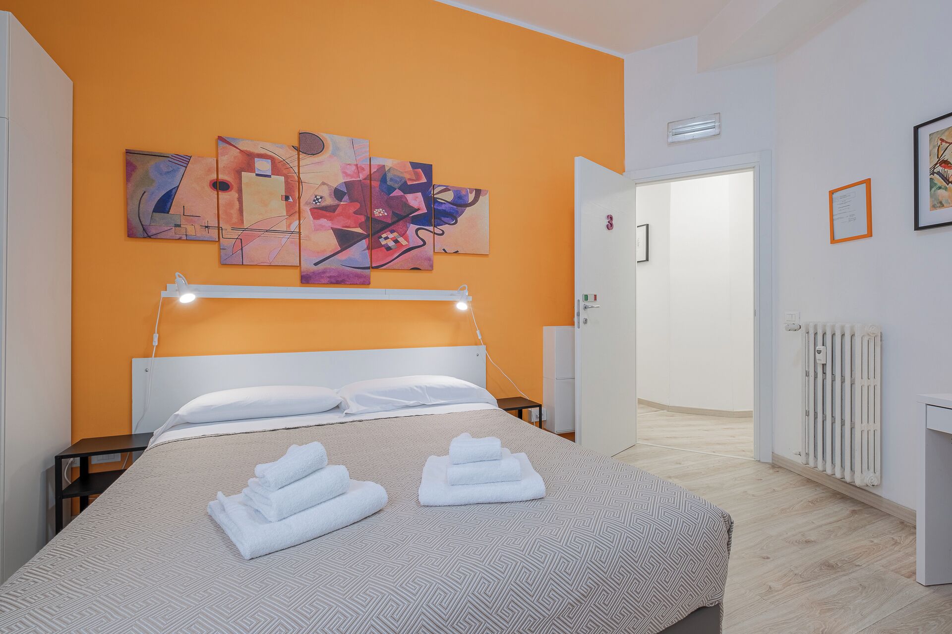 A hotel room with a bed, a nightstand, a wall-mounted lamp, and a door leading to another room.