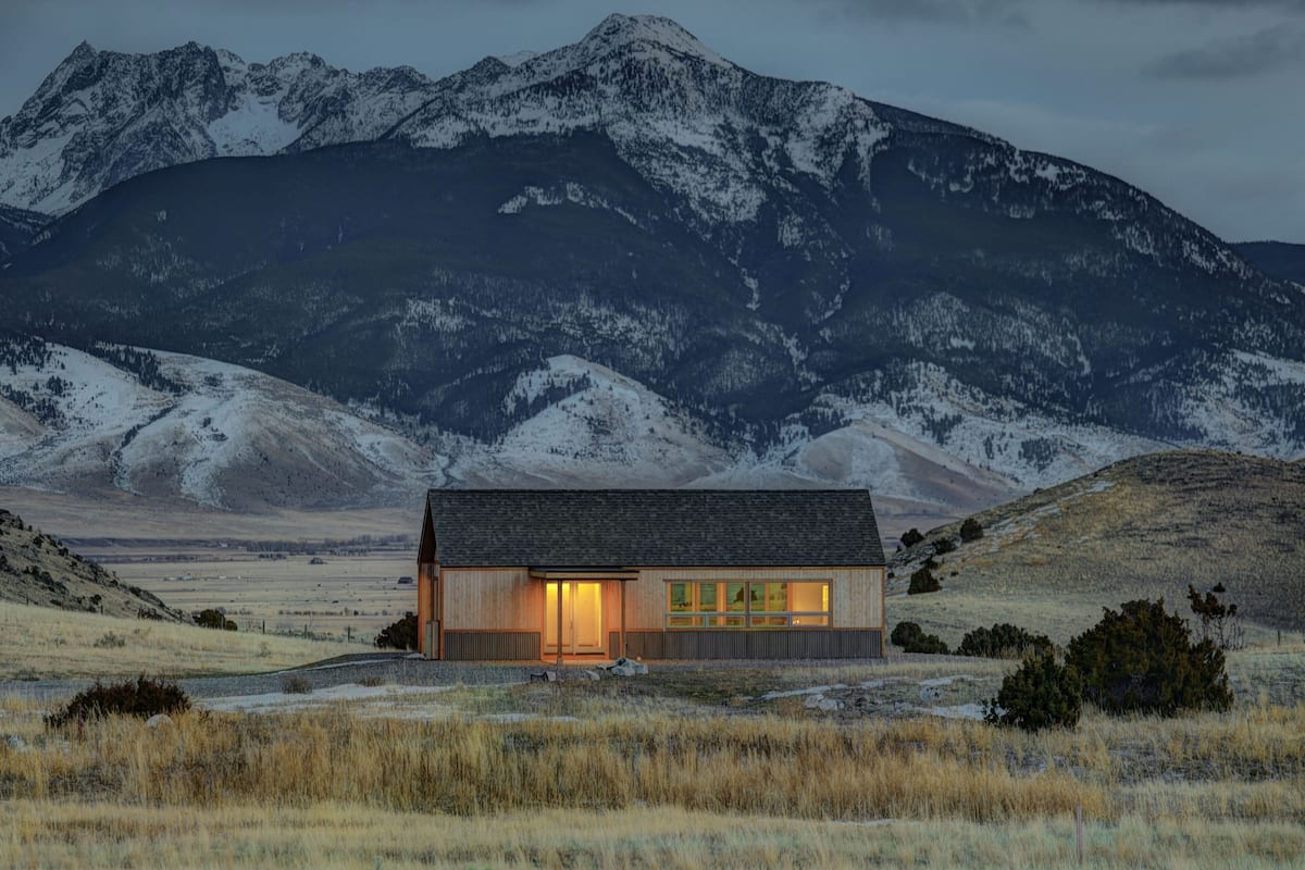 A modern cabin with large windows, situated in a mountainous area with snow-capped peaks in the background.
