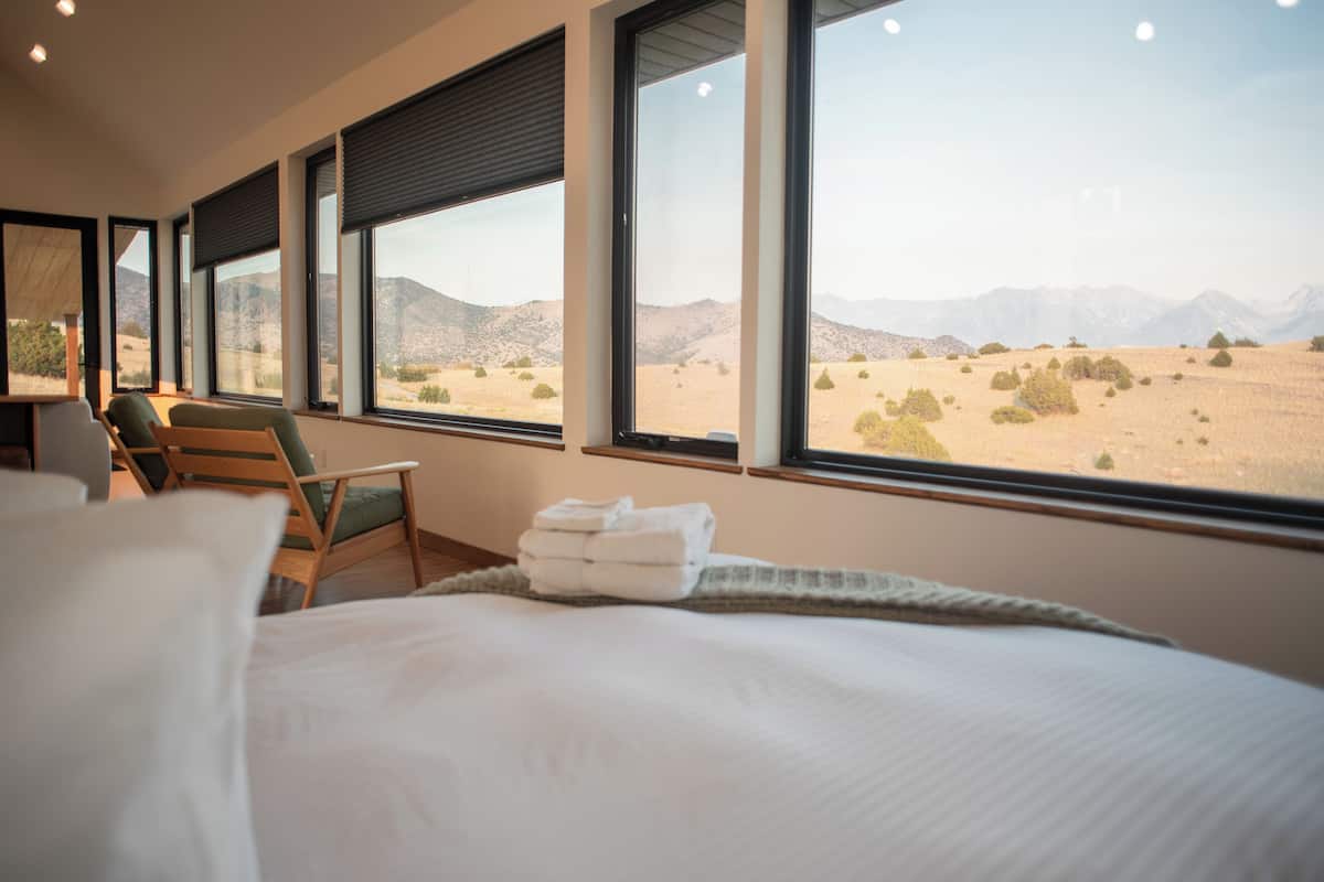 A hotel room with a bed, a chair, and large windows offering a desert view.