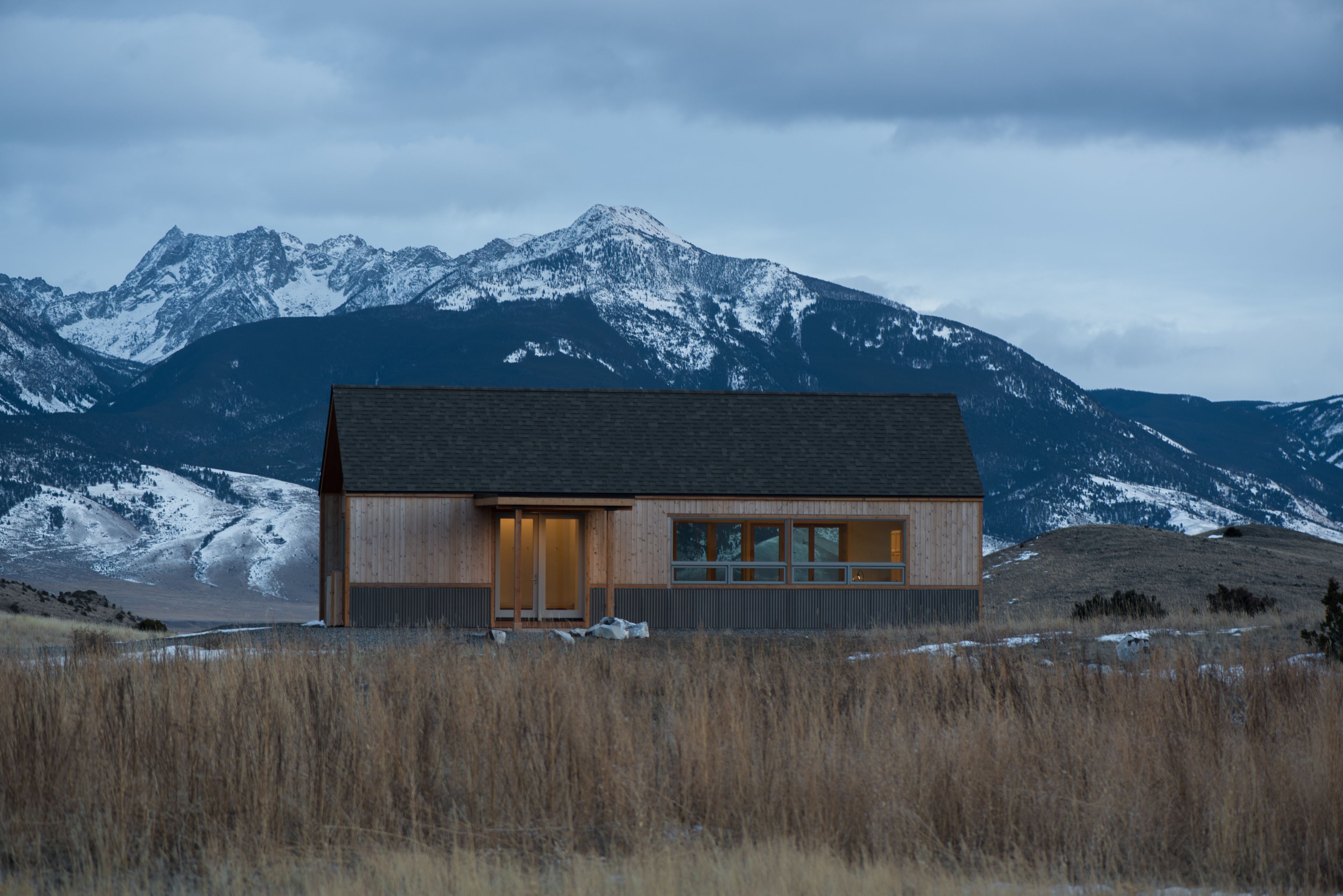 A modern wooden cabin with large windows, set against a backdrop of snow-capped mountains.