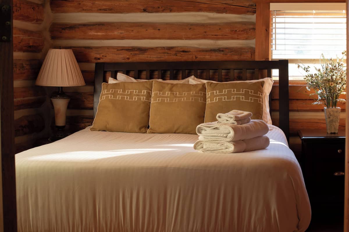 A neatly made bed with pillows and folded towels, a bedside lamp, and a vase with flowers on a wooden nightstand.