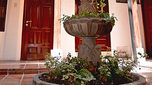 Une fontaine en pierre à plusieurs niveaux, agrémentée de plantes, installée dans une cour intérieure avec un balcon en bois et une porte rouge.