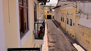 Une rue étroite, bordée de bâtiments des deux côtés, avec des balcons ornés de rampes en fer forgé et de jardinières.