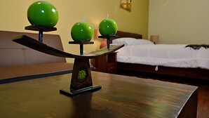 Une table en bois sur laquelle est posée une balance avec trois pommes vertes. En arrière-plan, on aperçoit un lit avec une literie blanche et une tête de lit marron.