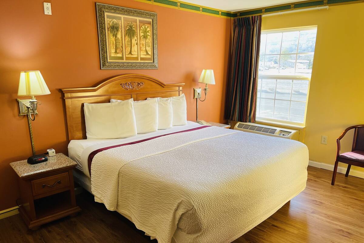 Una habitación de hotel con una cama grande, dos lámparas de noche, una mesita de noche, una silla, una ventana con cortinas y un cuadro enmarcado en la pared.