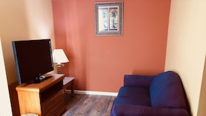 A living room with a flat-screen TV on a wooden stand, a small table with a lamp, a purple sofa, and a framed picture on the wall. - Mountain Inn & Suites Dunlap (Dunlap)