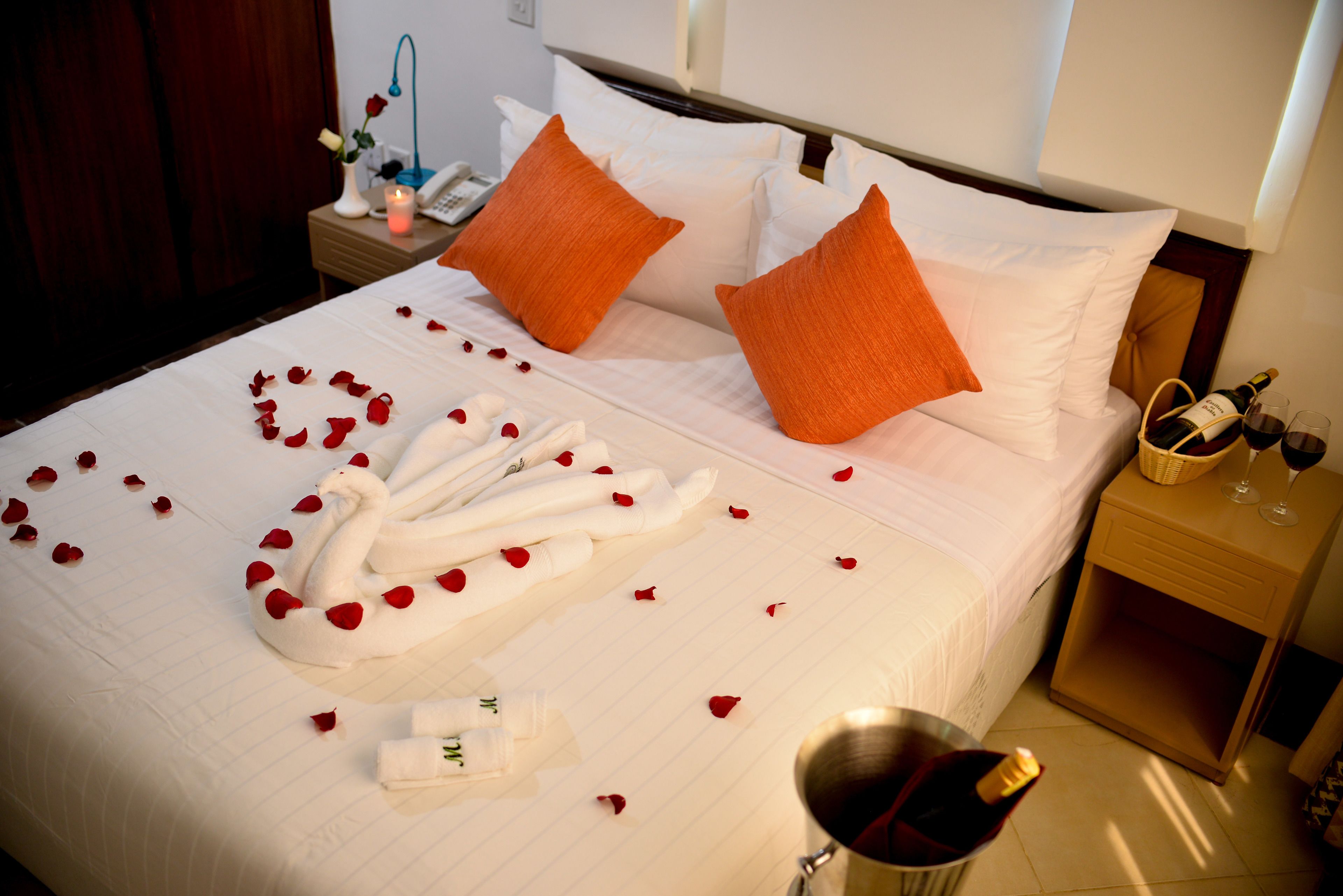 A neatly made bed with rose petals, a swan-shaped towel, and a bottle of champagne.