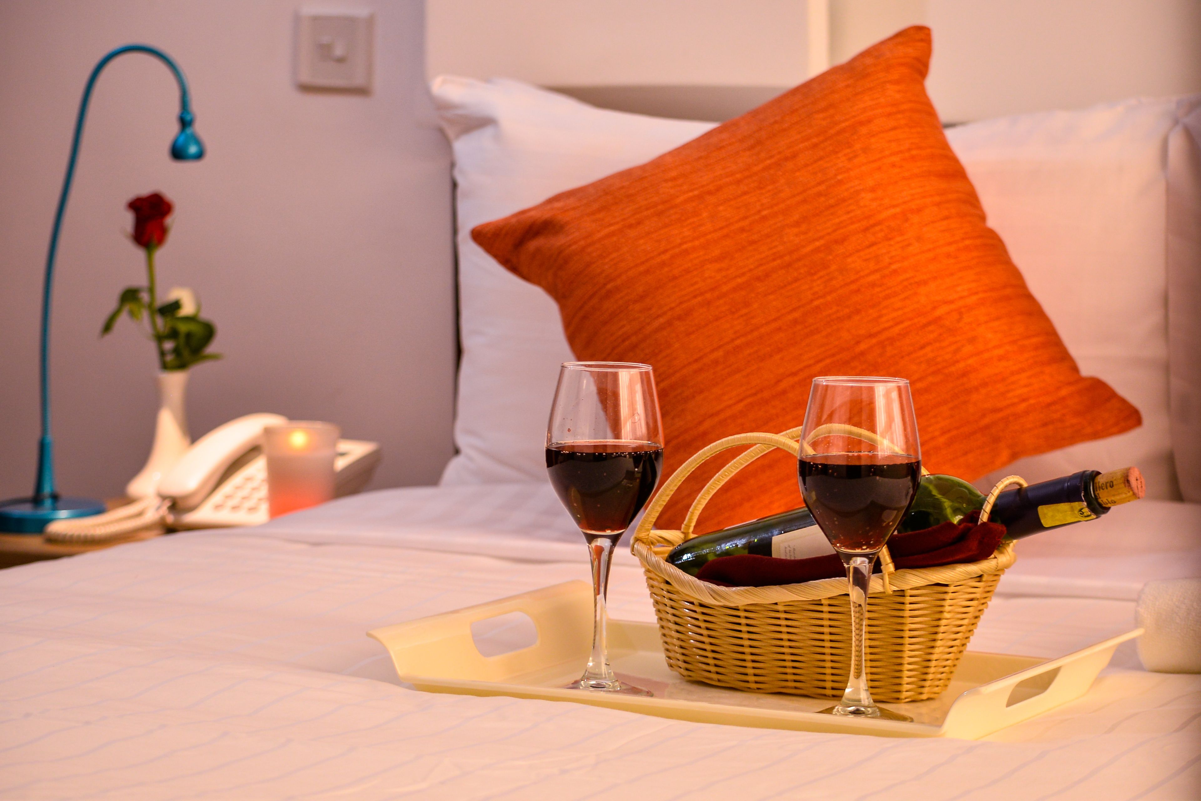 A neatly made bed with white bedding, a red rose in a vase, two glasses of red wine, and a bottle of wine in a wicker basket.