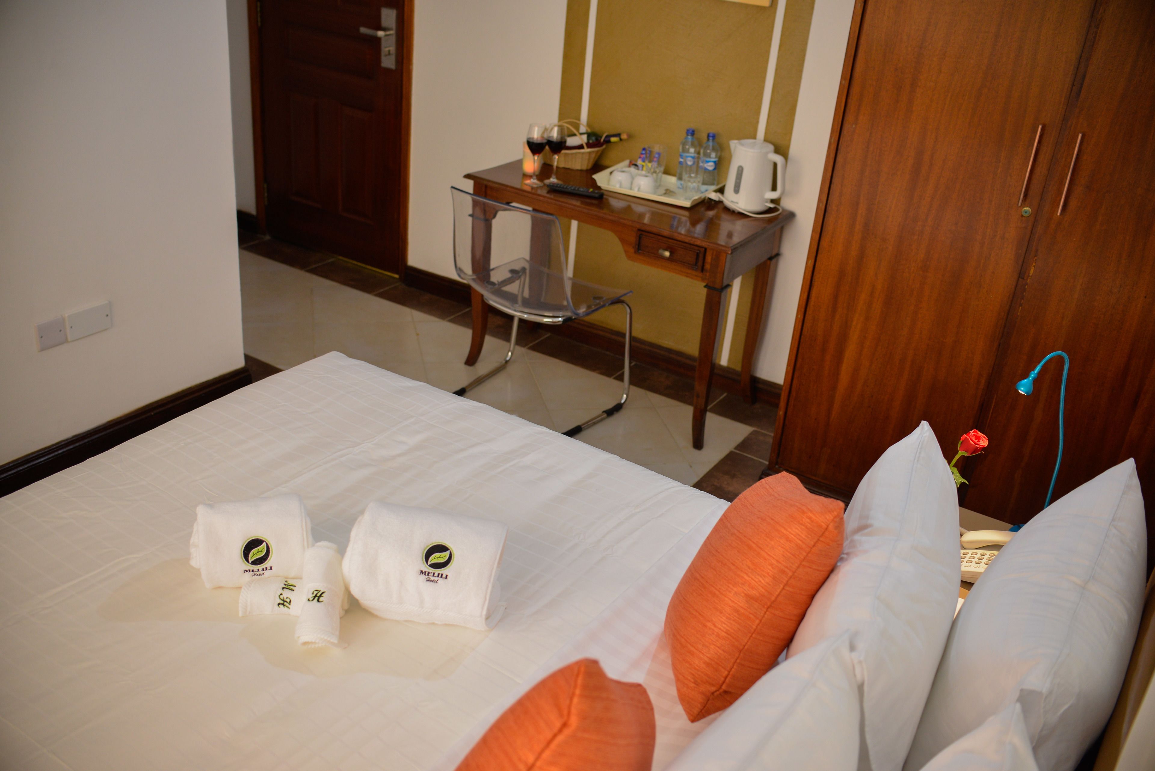 A hotel room with a bed, pillows, a desk with a chair, a nightstand, and a door.
