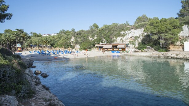 A beach with clear water, a building with a roof, and palm trees. - Pierre & Vacances Menorca Cala Blanes (Ciutadella de Menorca)