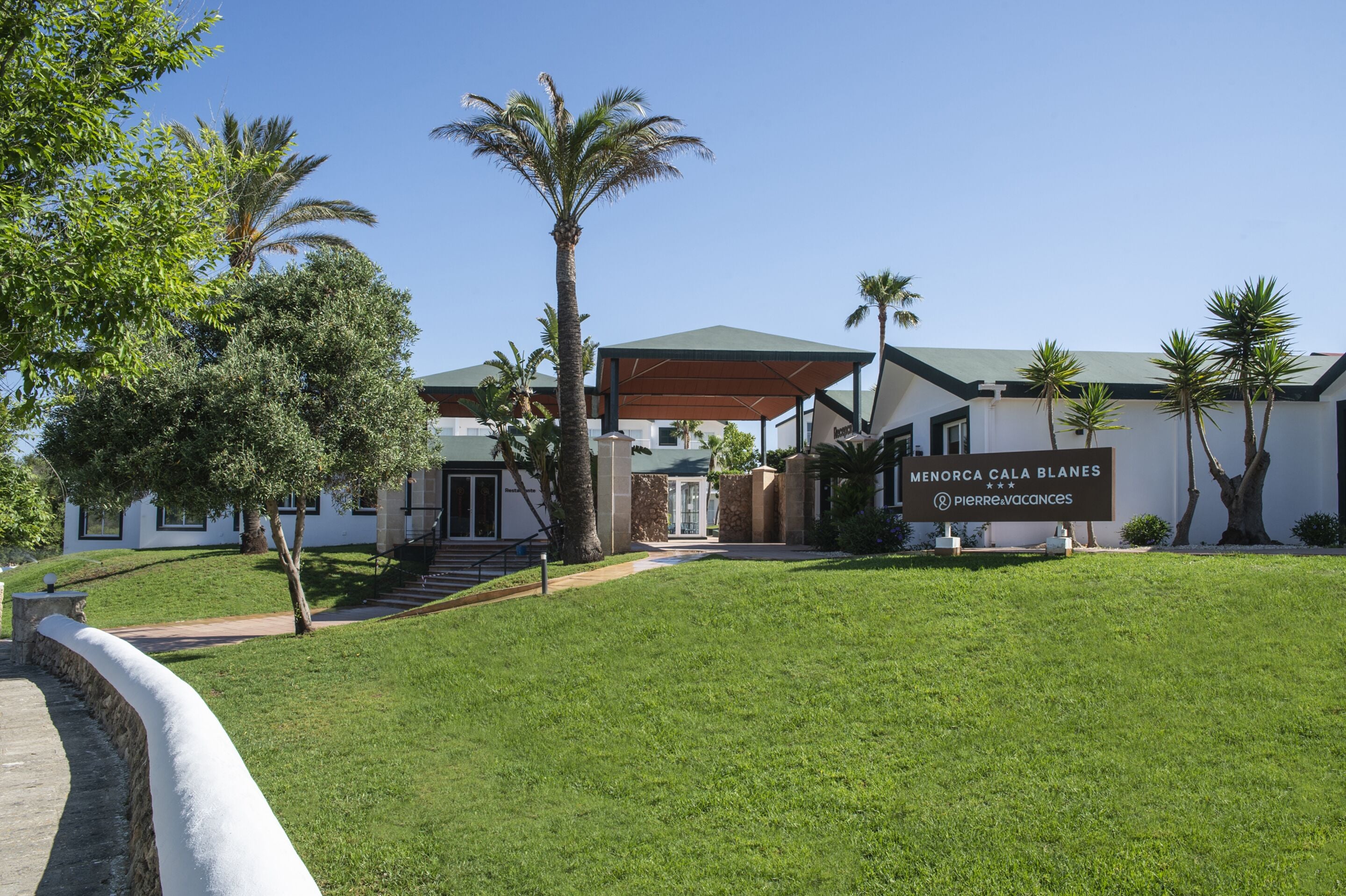 A modern hotel with a covered entrance, palm trees, and a sign reading "Menorca Cala Blanes."