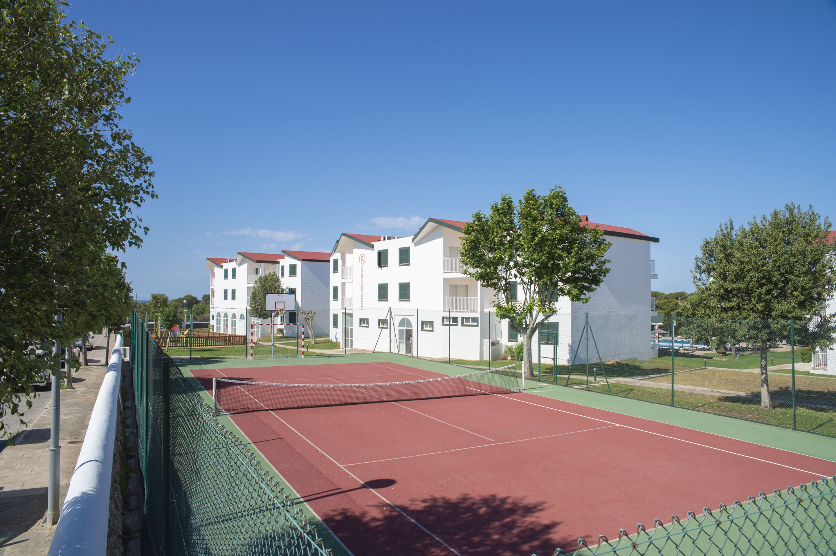 Una cancha de tenis con valla verde, rodeada de árboles y edificios con techos rojos.