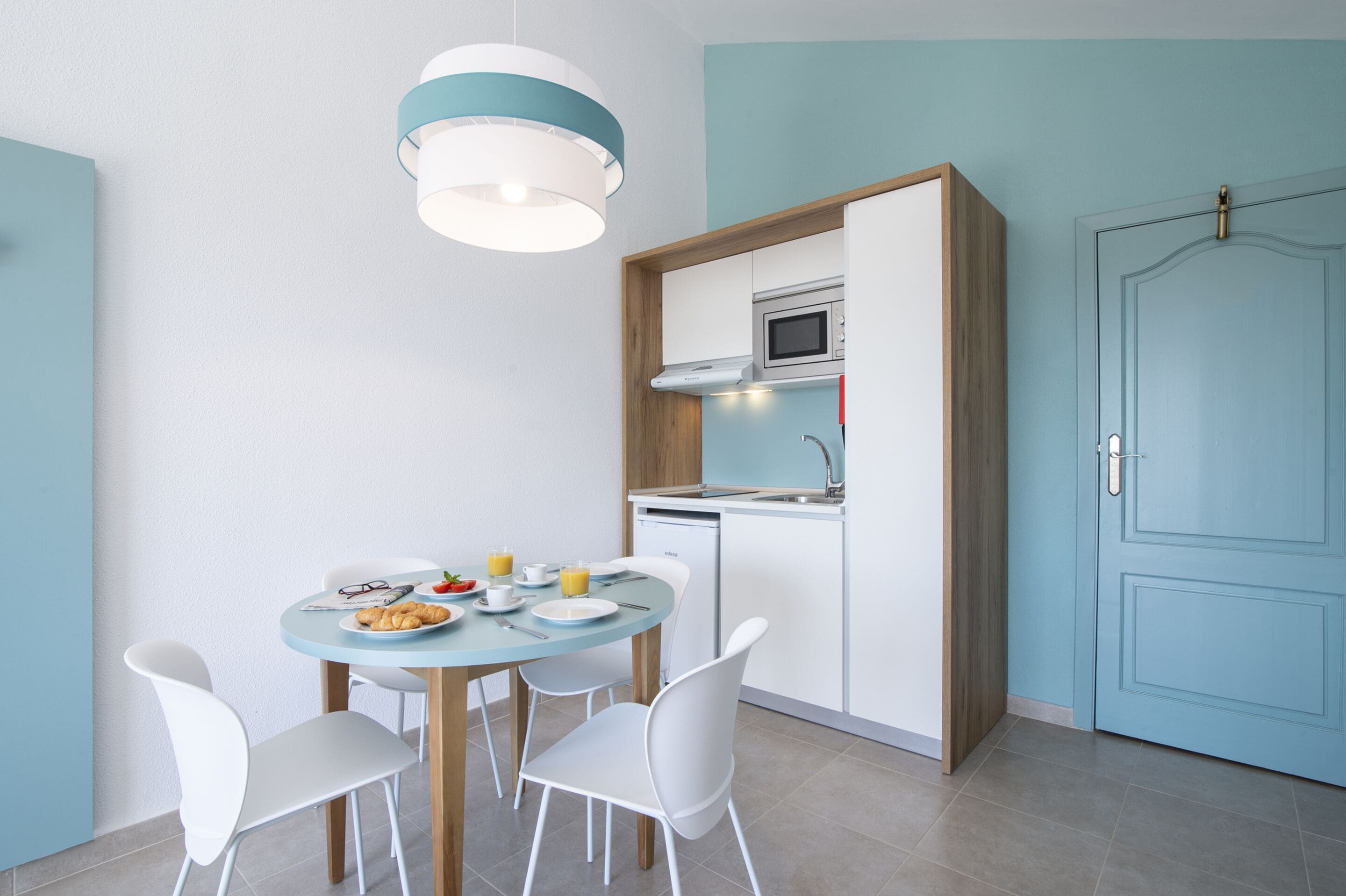 A modern kitchen with a dining area, featuring a round table with white chairs, a blue door, and a hanging light fixture.