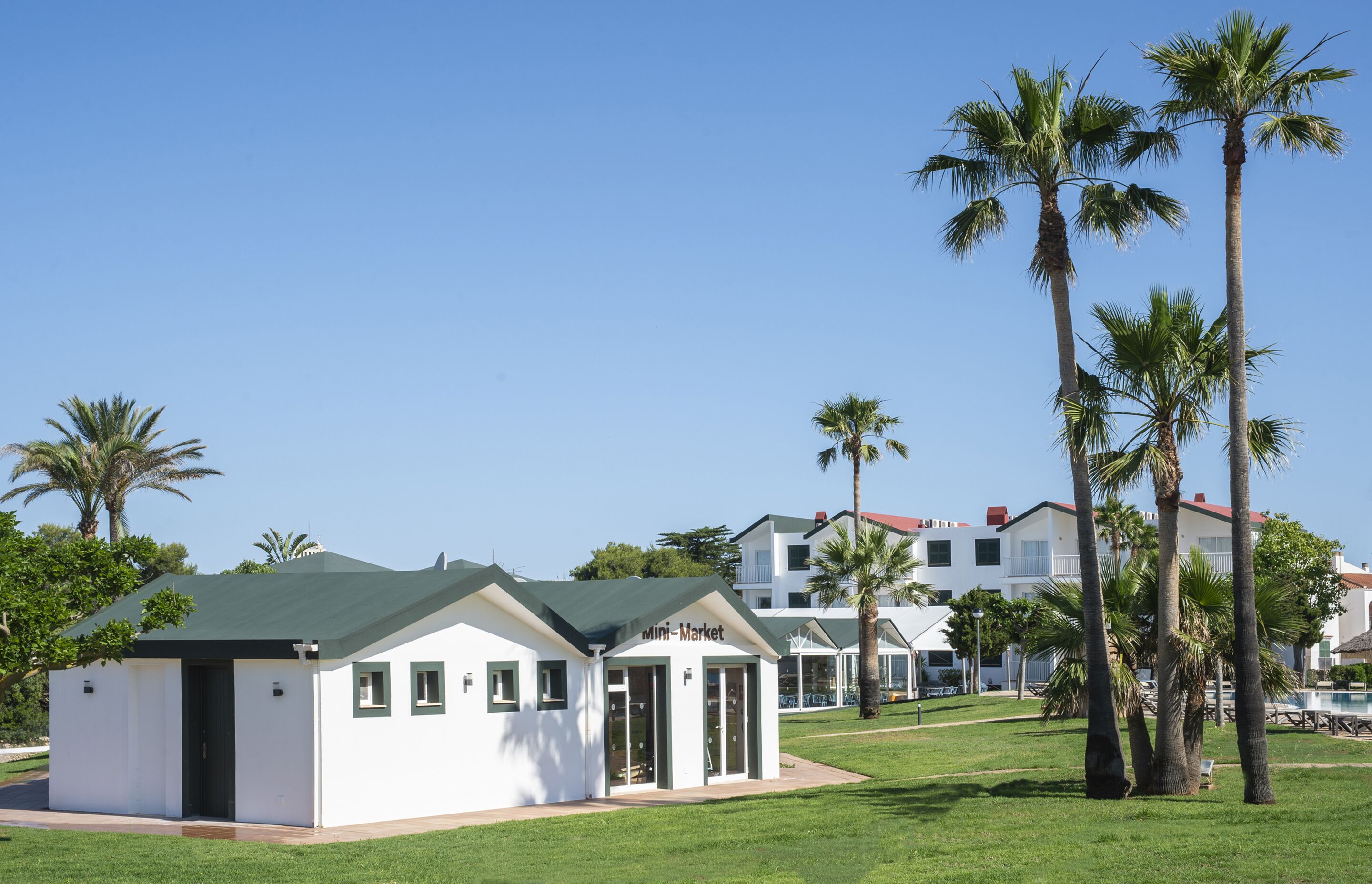 A white building with a green roof labeled "Mini-Market" in front of a row of white houses with red roofs and palm trees.