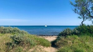 Ein Küstenpfad führt zu einem Sandstrand mit freiem Blick auf das Meer und einem Segelboot in der Ferne.