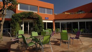 An outdoor seating area with green chairs and tables, a reception desk, and a building with an orange facade. - Relaxed Beachside Living (LE CAP D'AGDE)