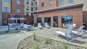An outdoor patio area with seating and tables, surrounded by a brick building with large windows.
