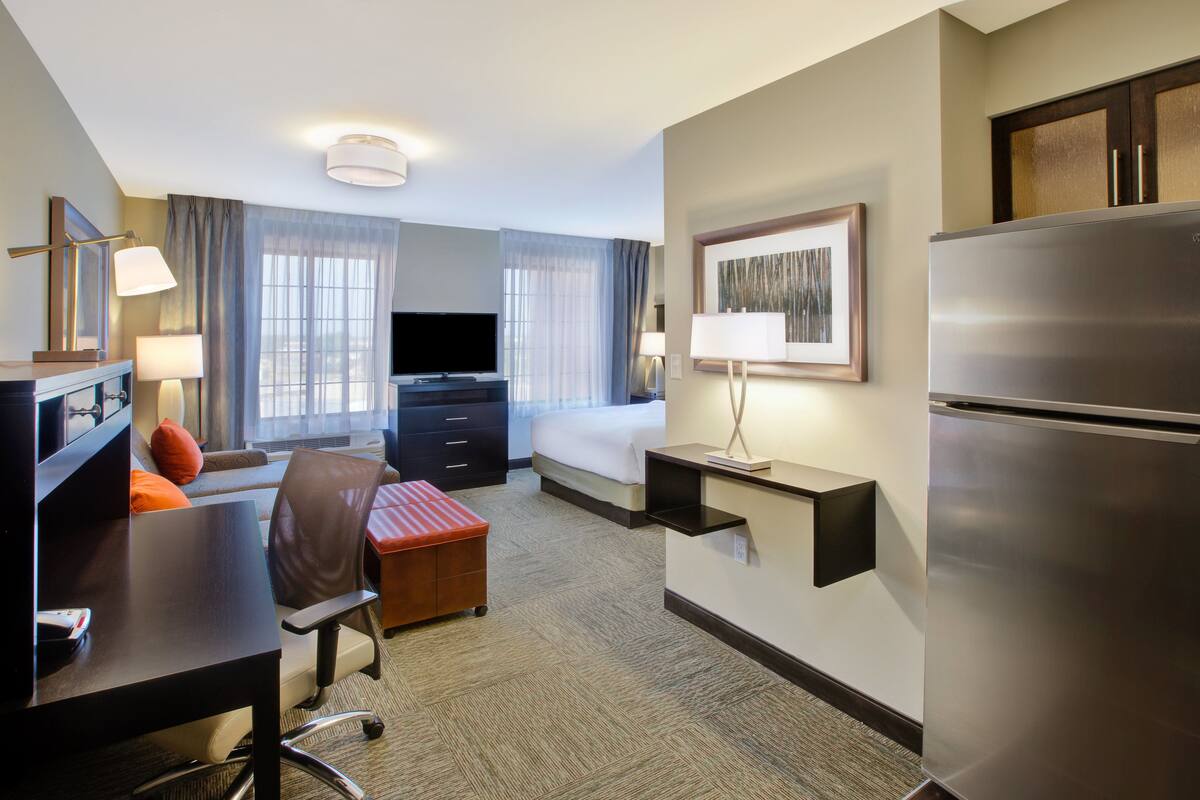 A hotel room with a bed, desk, chair, television, refrigerator, and a small table with a lamp.