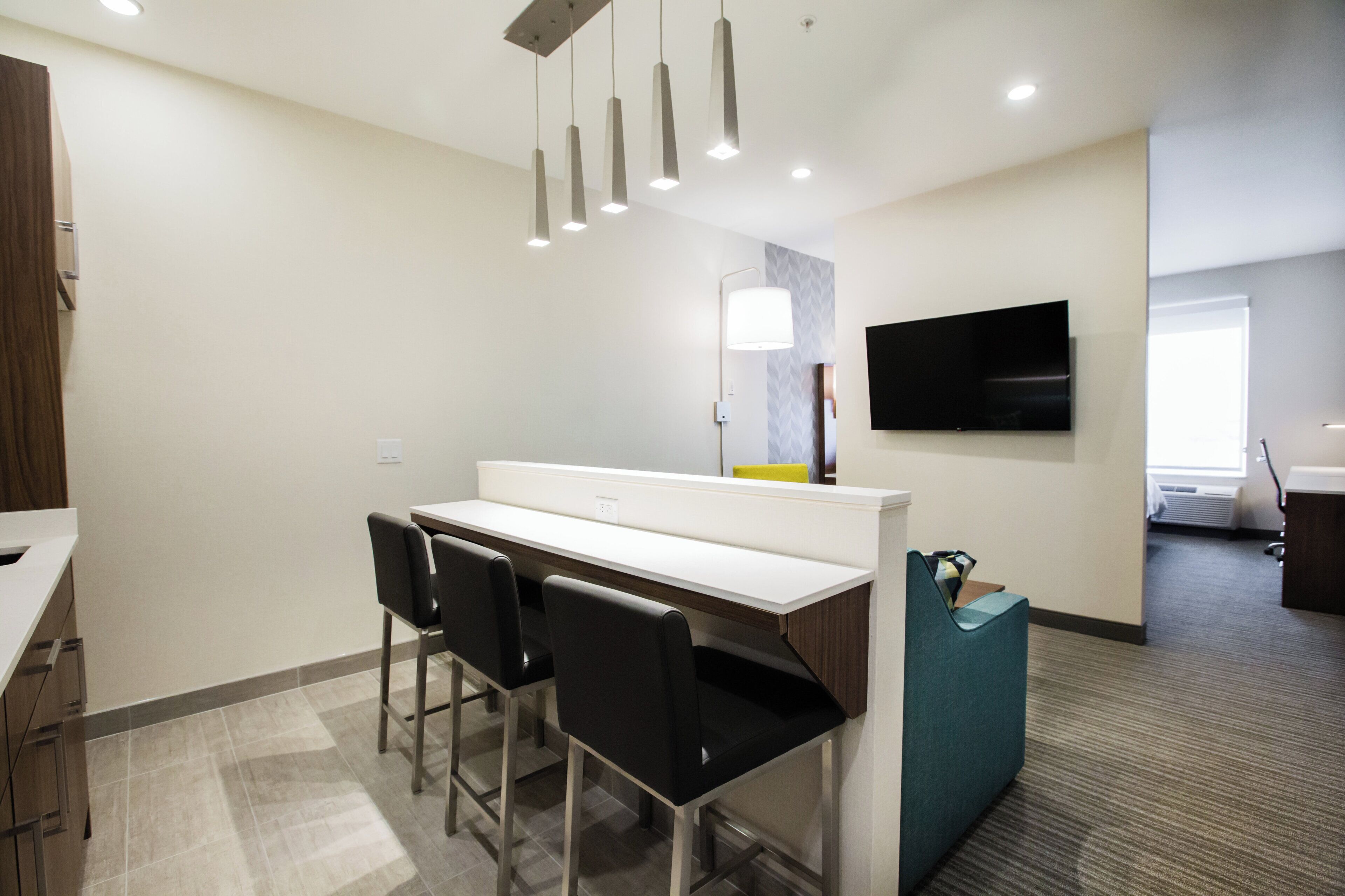 A modern hotel room with a kitchenette, a flat-screen TV mounted on the wall, and a seating area with chairs.