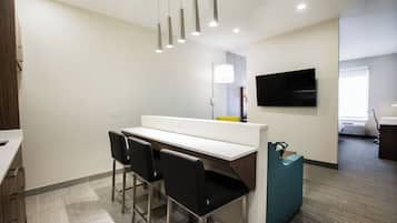 A modern hotel room with a kitchenette, a flat-screen TV mounted on the wall, and a seating area with chairs.