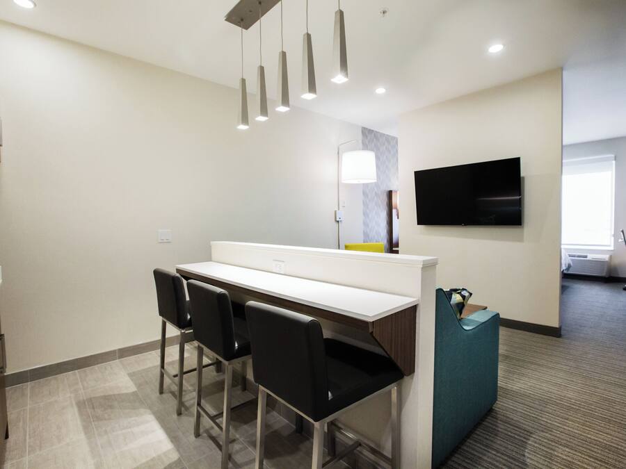 A modern hotel room with a kitchenette, a flat-screen TV mounted on the wall, and a seating area with chairs.
