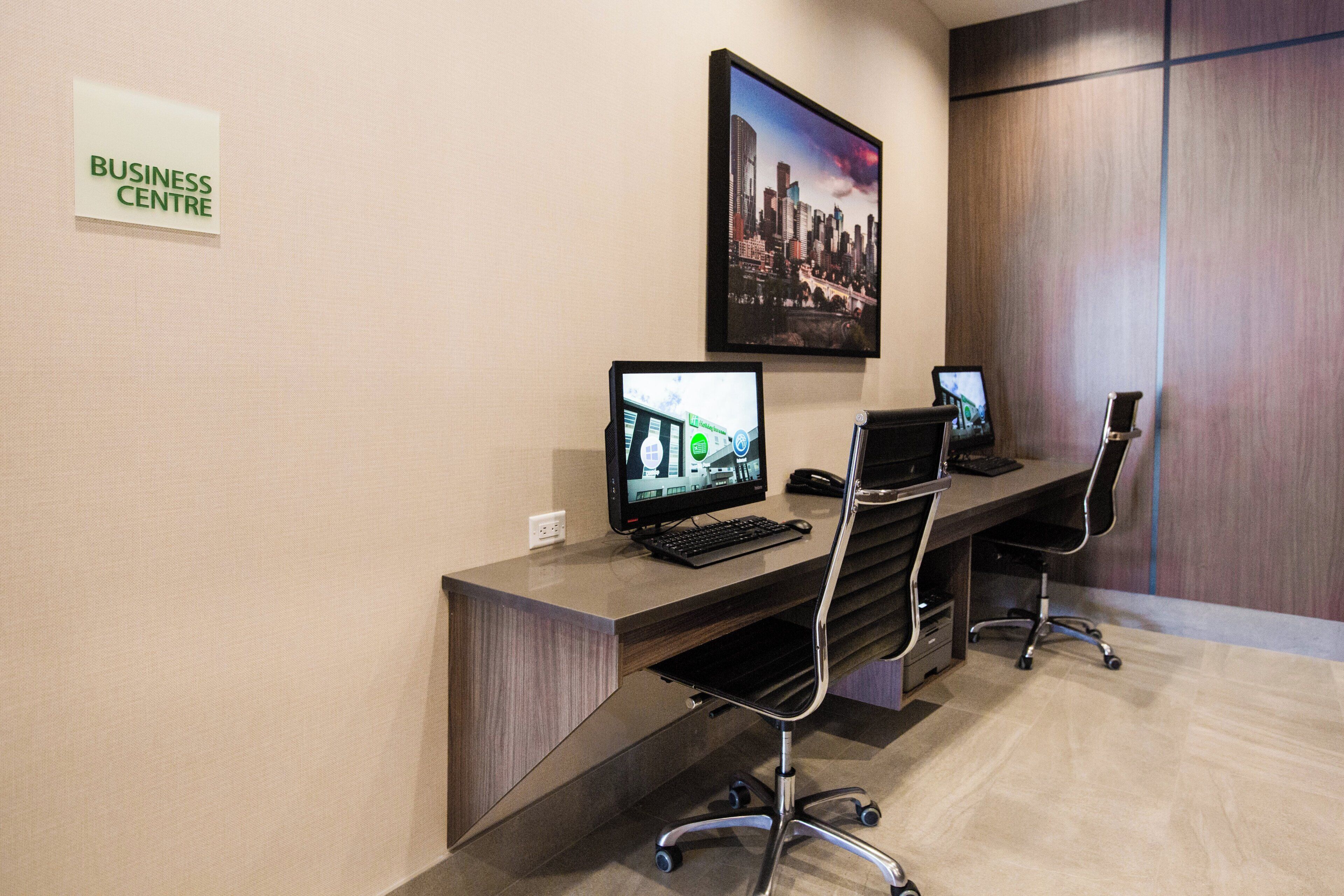 Un centro de negocios con dos estaciones de trabajo de computadora, una silla e imagen de un paisaje urbano en la pared.