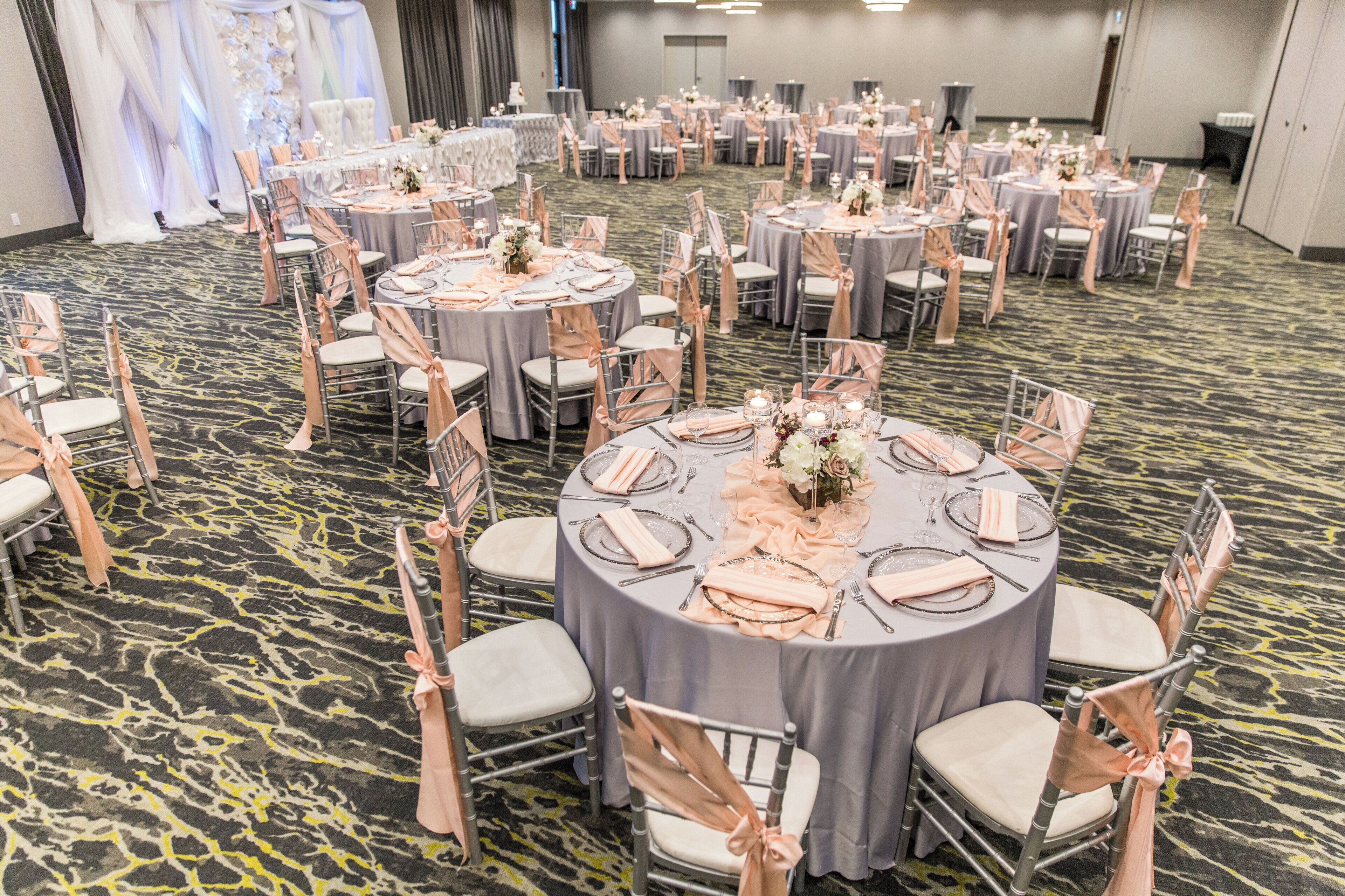 Une salle de banquet avec des tables rondes disposées pour des événements, dotée de chaises recouvertes d’un tissu de couleur claire et d’un tapis à motifs.