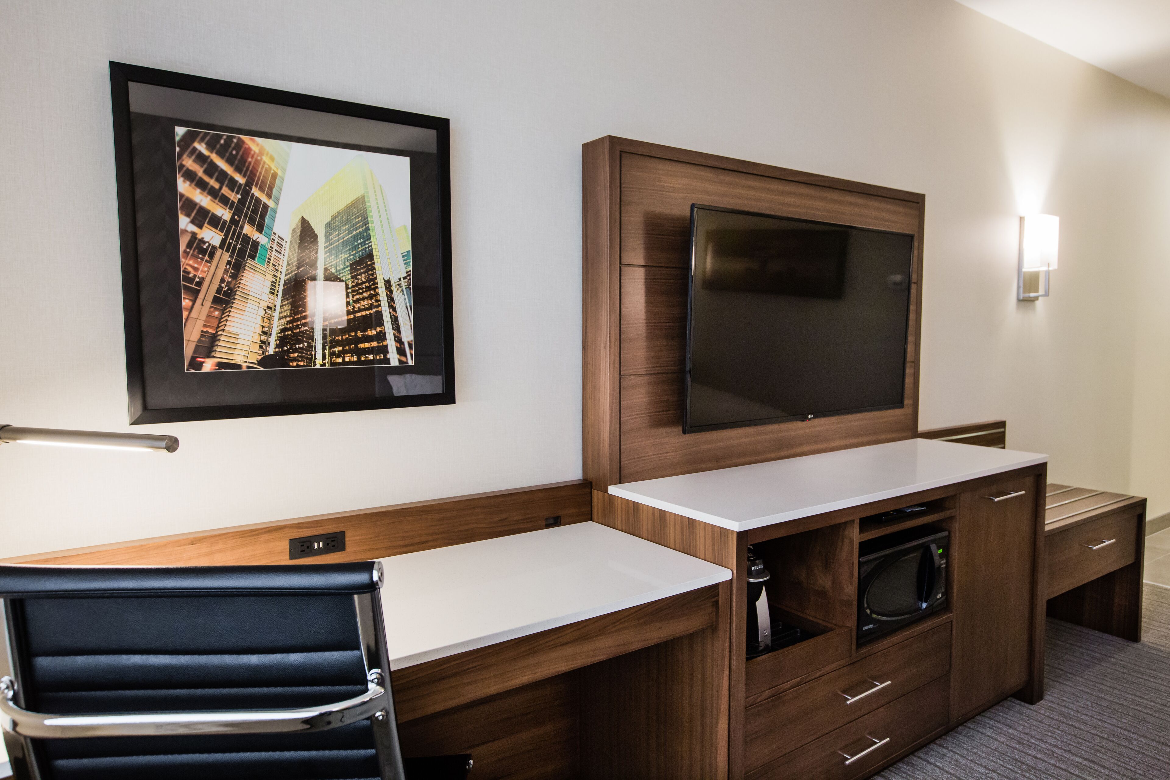 A hotel room with a wooden desk, a flat-screen TV, a framed cityscape photo, and a microwave.