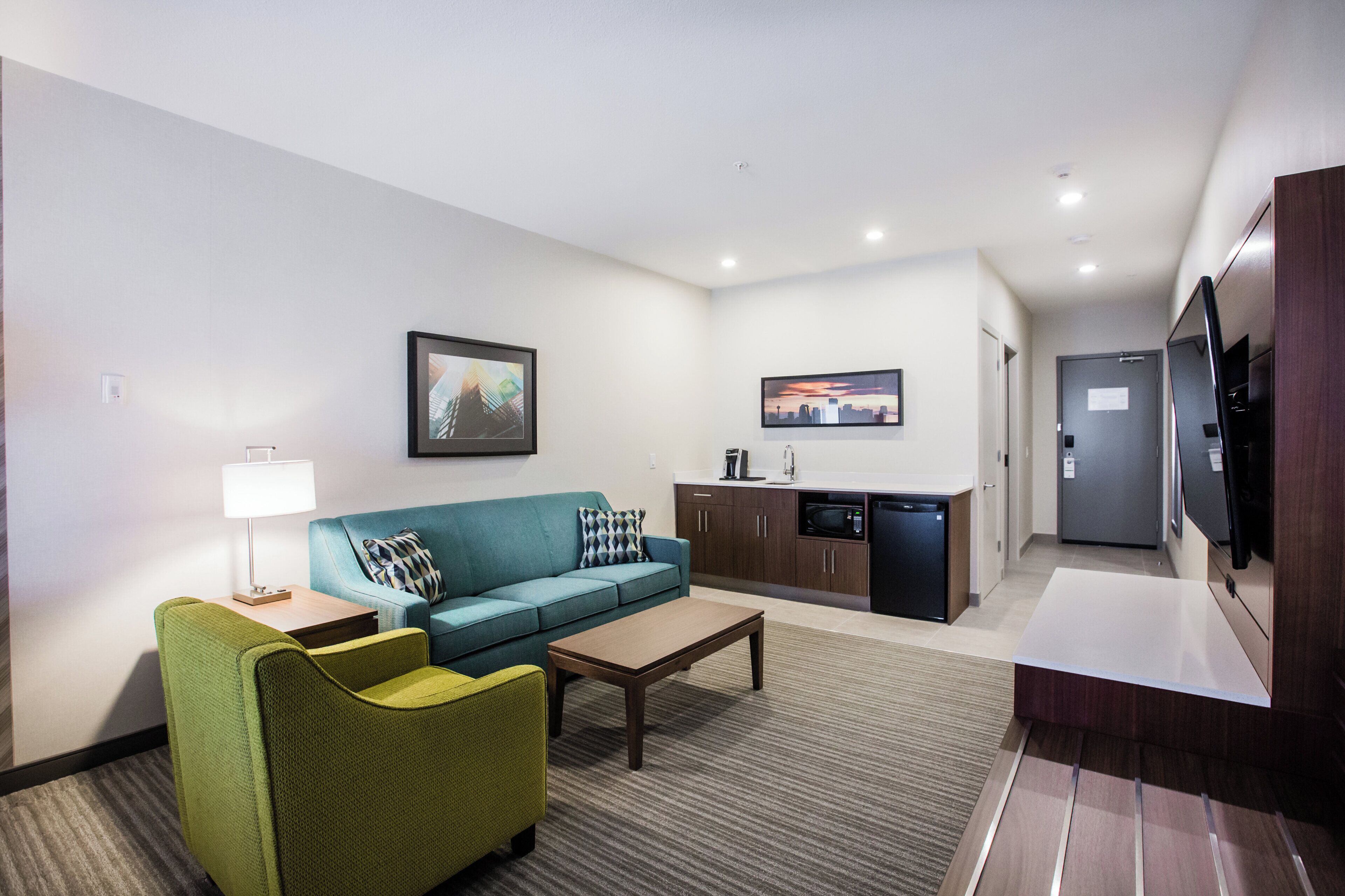 A compact hotel room with a sofa, a small coffee table, a kitchenette, and a television.