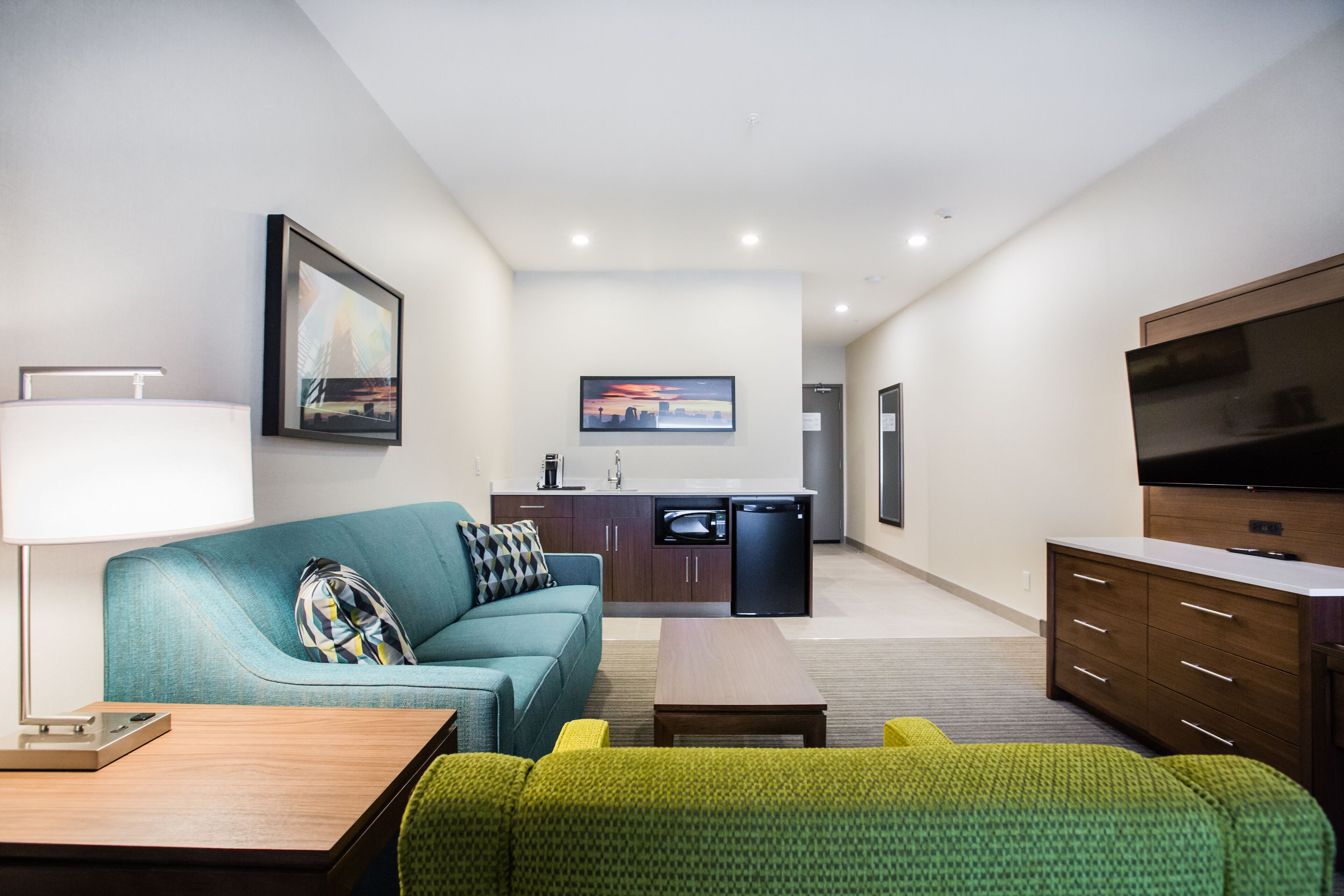 A modern hotel room with a sofa, a small table, a TV, a kitchenette, and a large window.