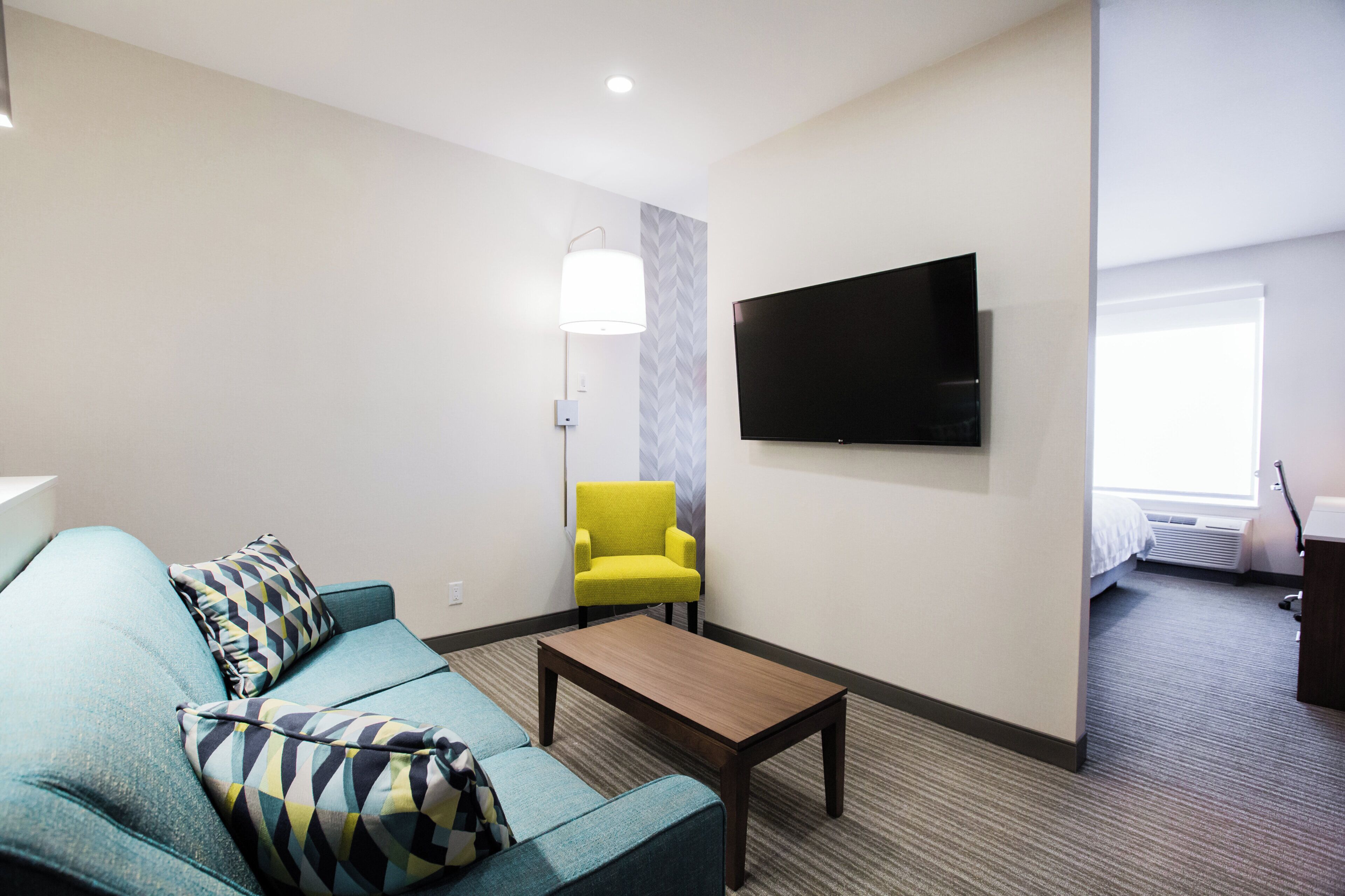 A hotel room with a sofa, a yellow armchair, a coffee table, a flat-screen TV mounted on the wall, and a window with a view.