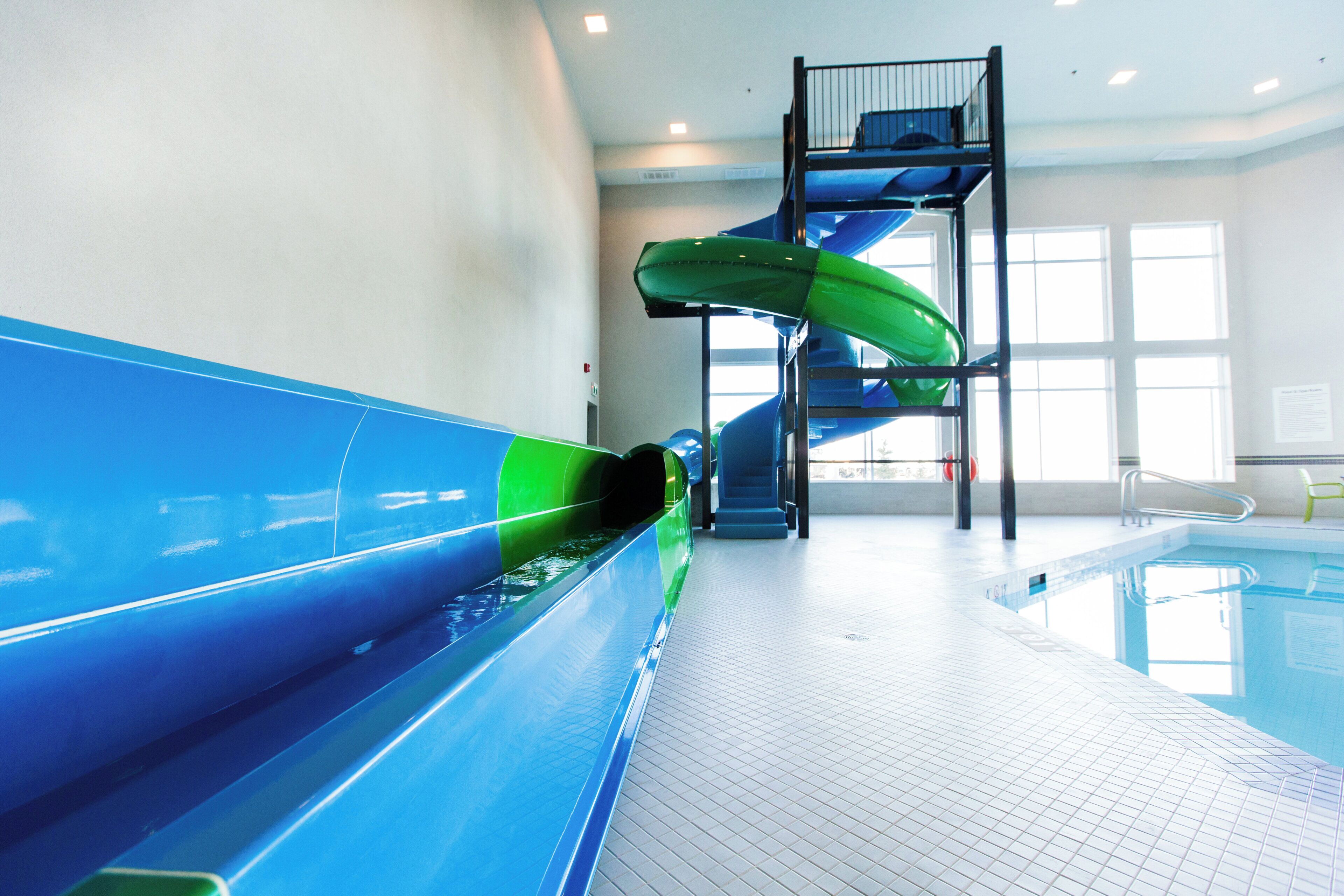 Eine Indoor-Wasserrutsche mit blau-grünen Farbtönen, ein Schwimmbecken und eine Treppe zu einer erhöhten Plattform.