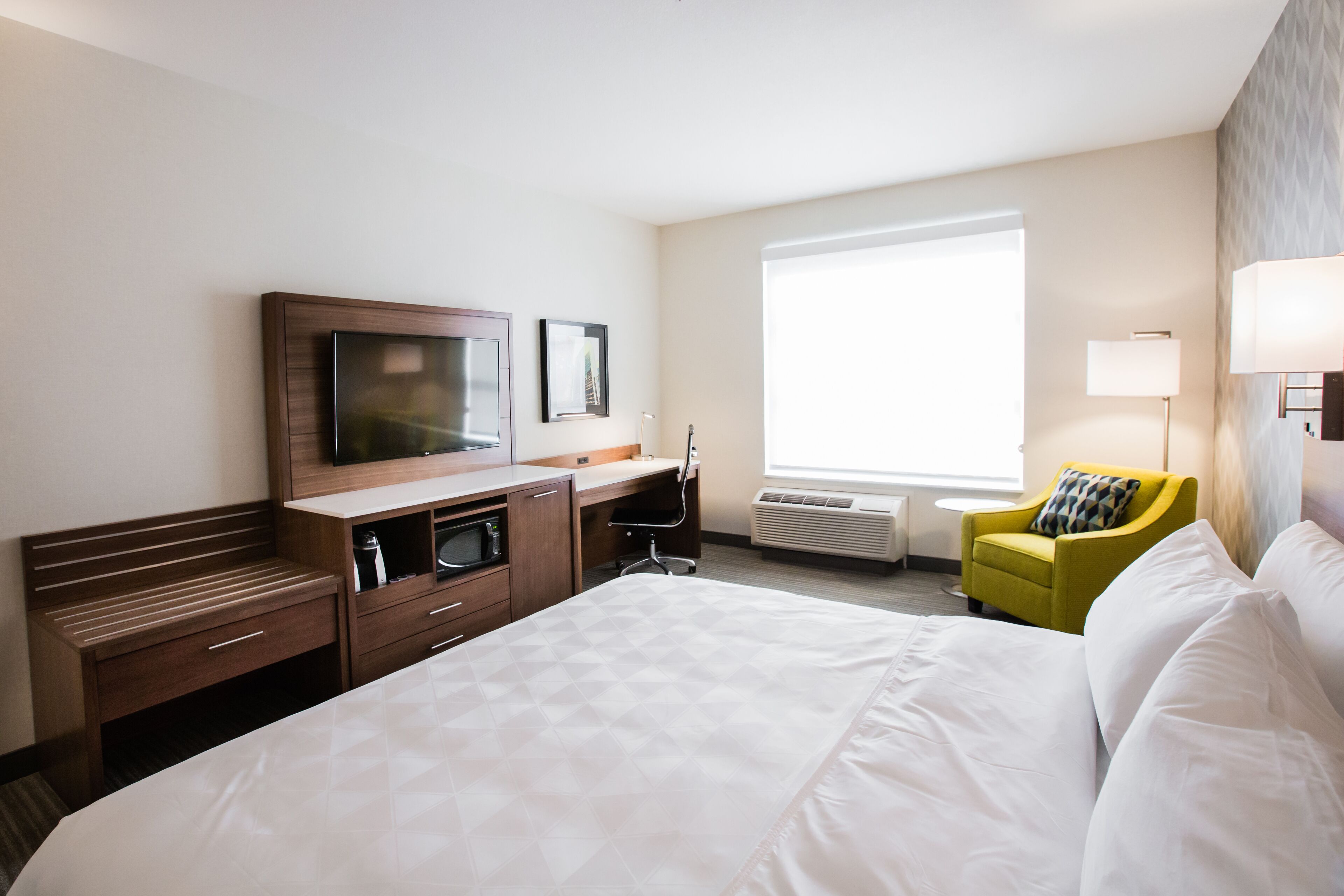 A hotel room with a large bed, a desk with a chair, a TV, a window, and a lamp.
