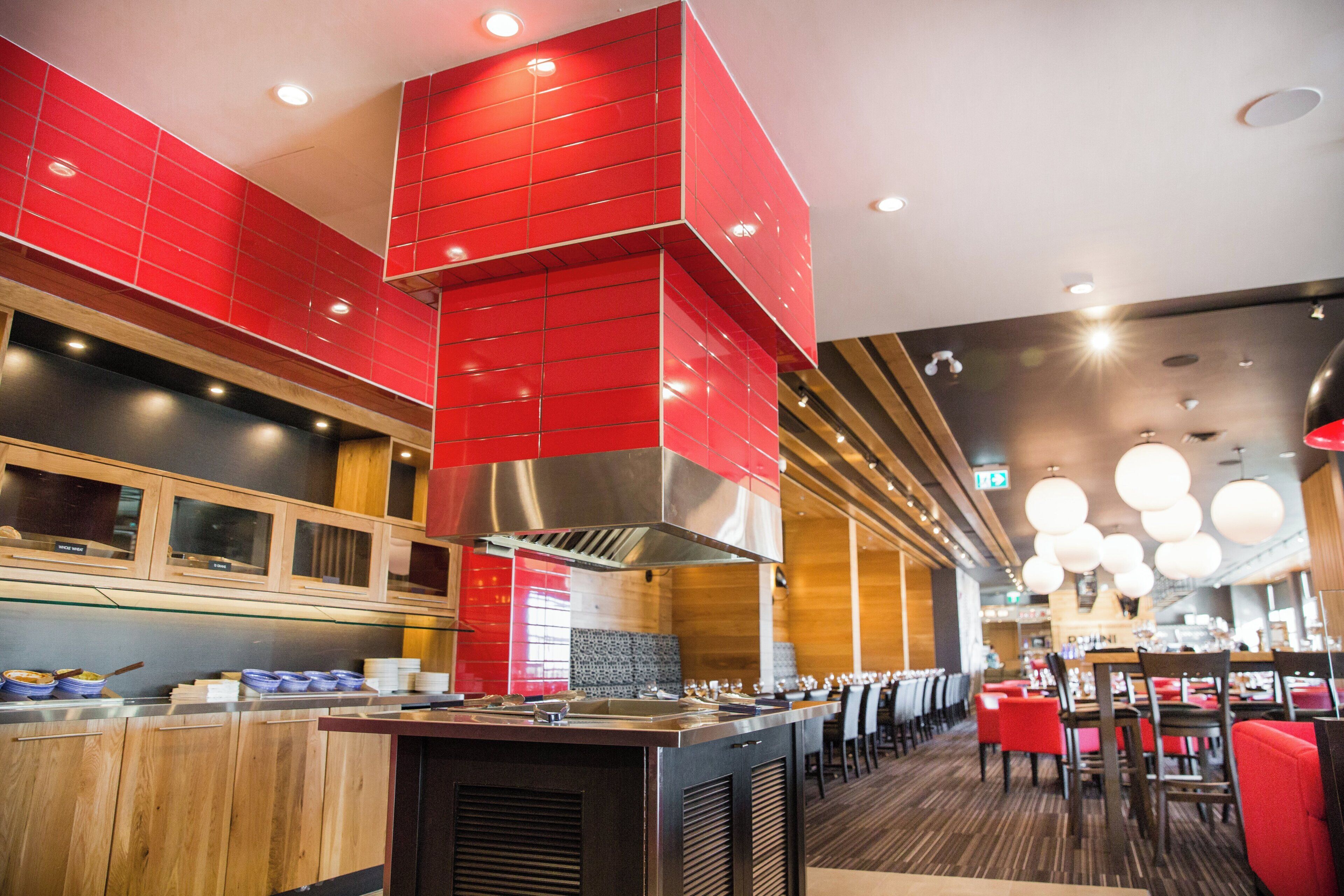Interior moderno de restaurante con detalles rojos, paneles de madera y una isla central con equipo de cocina.