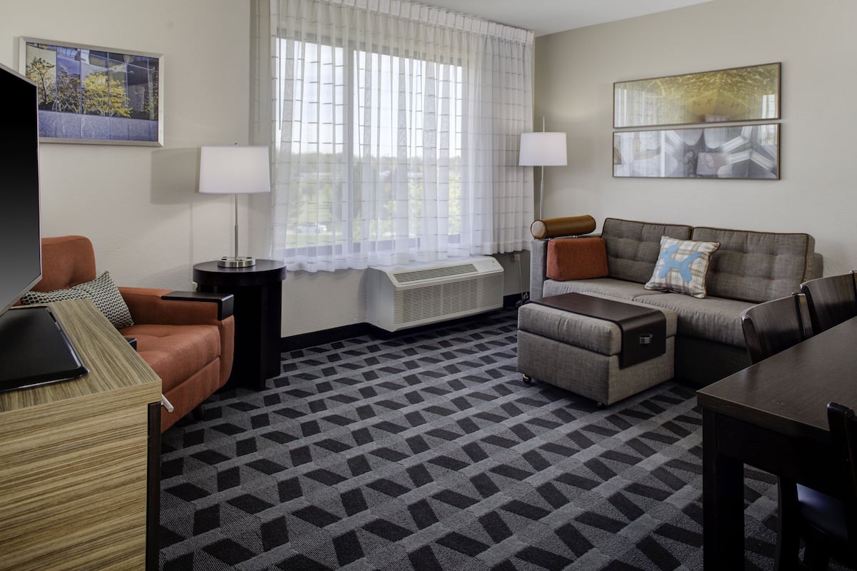 A modern hotel room with a sofa, armchair, and a small table. There is a window with sheer curtains and a radiator.