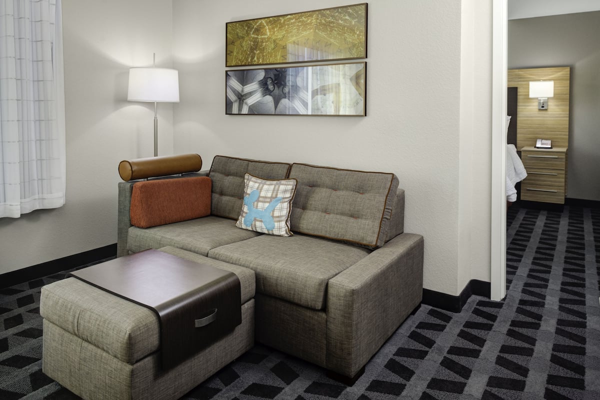 A hotel room with a sofa, a side table, a lamp, and a framed artwork on the wall.