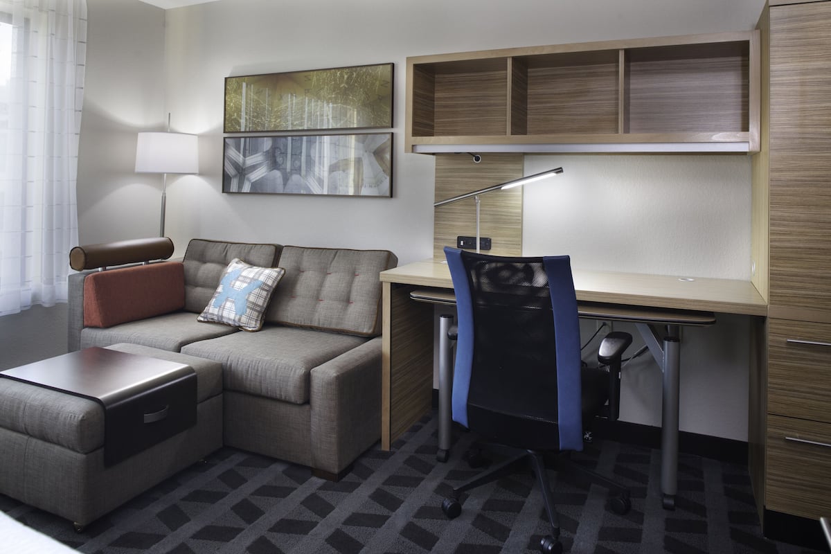 A modern hotel room with a sectional sofa, a desk, a chair, a lamp, and artwork on the walls.