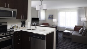 A modern kitchen with a microwave, stove, and dishwasher. There is a dining area with a table and chairs, and a living room with a sofa and a lamp. - TownePlace Suites by Marriott Detroit Canton (Canton)