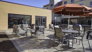 An outdoor patio area with multiple tables and chairs, a brick building in the background, and a large umbrella providing shade. - TownePlace Suites by Marriott Detroit Canton (Canton)