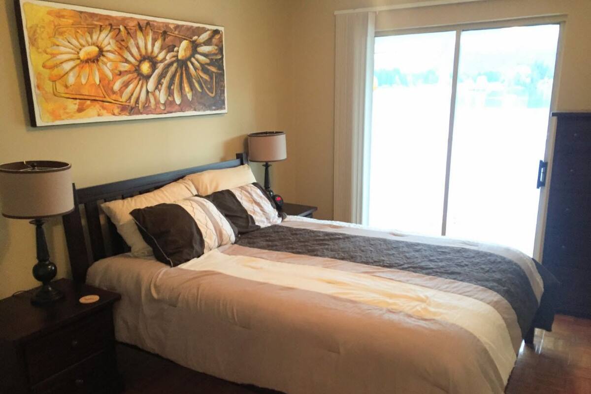A neatly made bed with a white and gray duvet, two bedside lamps, and a large window with a view of greenery.