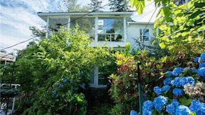 A two-story house with a visible front porch and a garden with various plants and blue flowers. - Leigh Lani Guest House (Victoria)