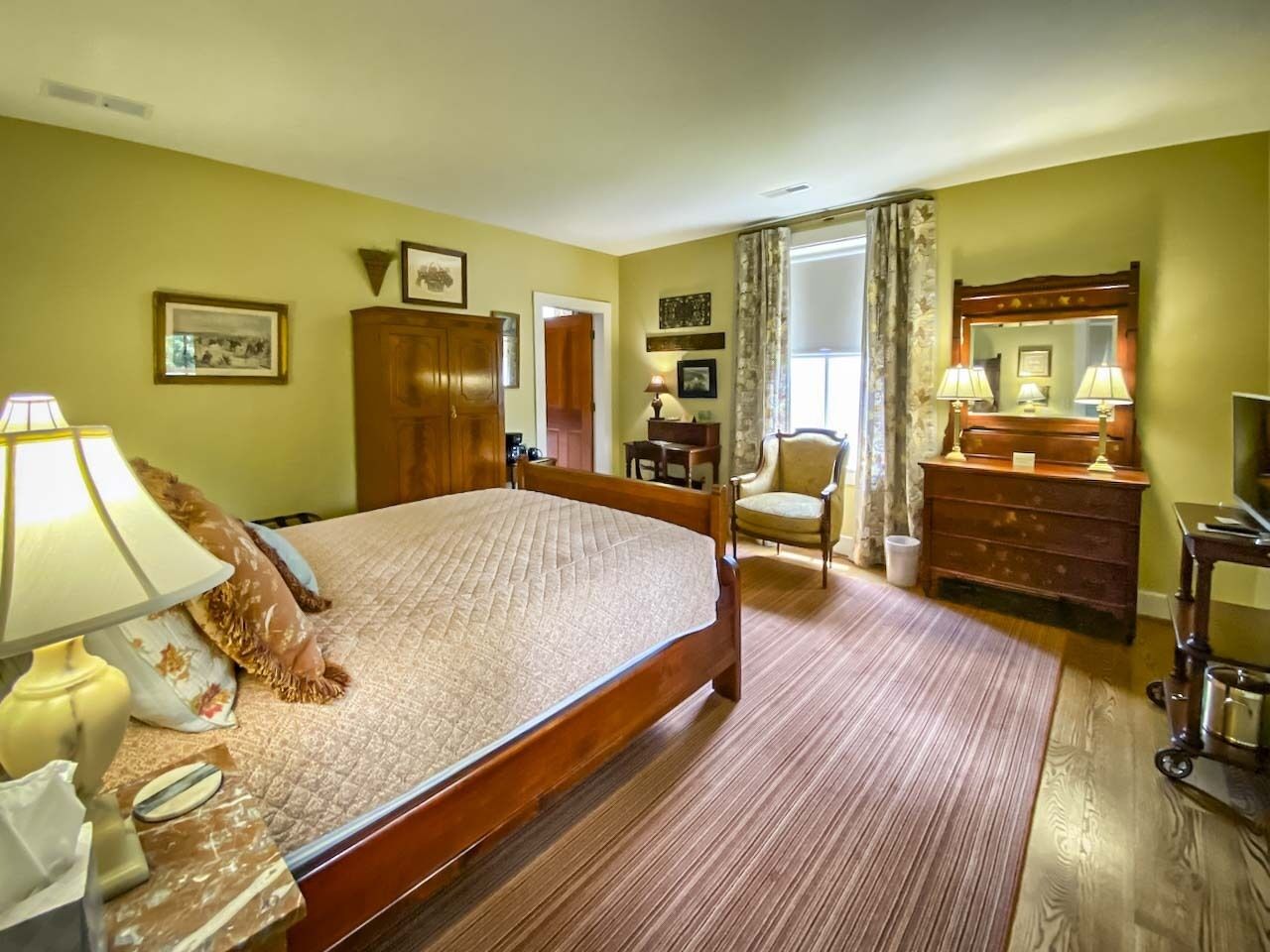 A hotel room with a large bed, a wooden dresser, a chair, and a desk with a lamp.