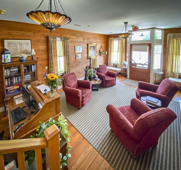 A cozy living room with wooden walls, a fireplace, and a large window. There are four red armchairs, a wooden coffee table, and a bookshelf filled with books. - Central Hotel (New Haven)