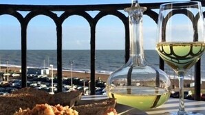 A table with a bottle of white wine, a glass of white wine, and a plate of food on a balcony overlooking the sea. - Moray Hastings (Hastings)