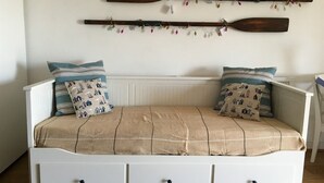 A white sofa with a beige blanket and patterned pillows, a wooden wall-mounted shelf with decorative items, and a white wall with a window. - Moray Hastings (Hastings)
