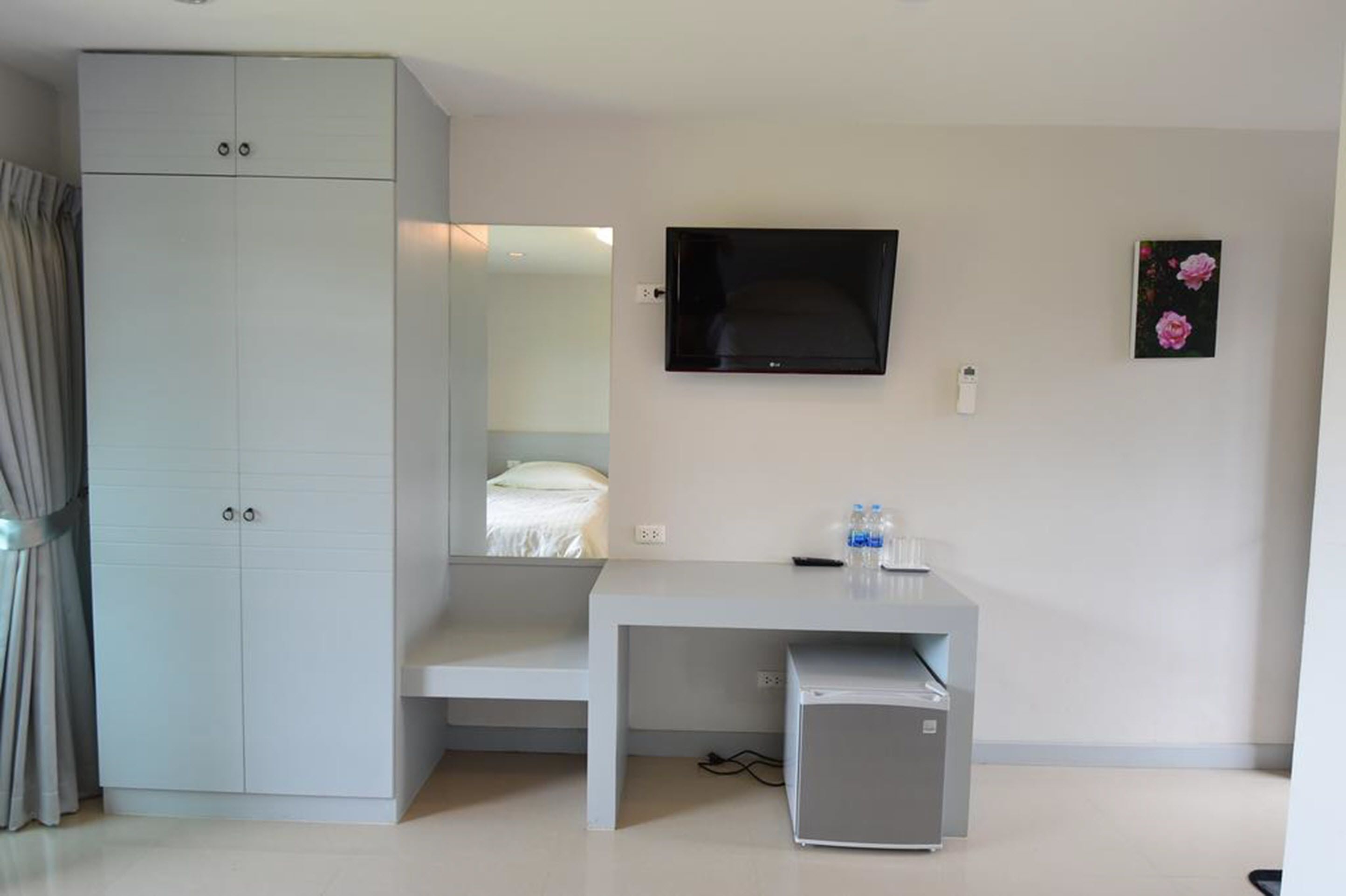 Habitació d'hotel amb un armari gris, una televisió de pantalla plana penjada a la paret, un taulell petit amb una ampolla d'aigua i un frigorífic petit.