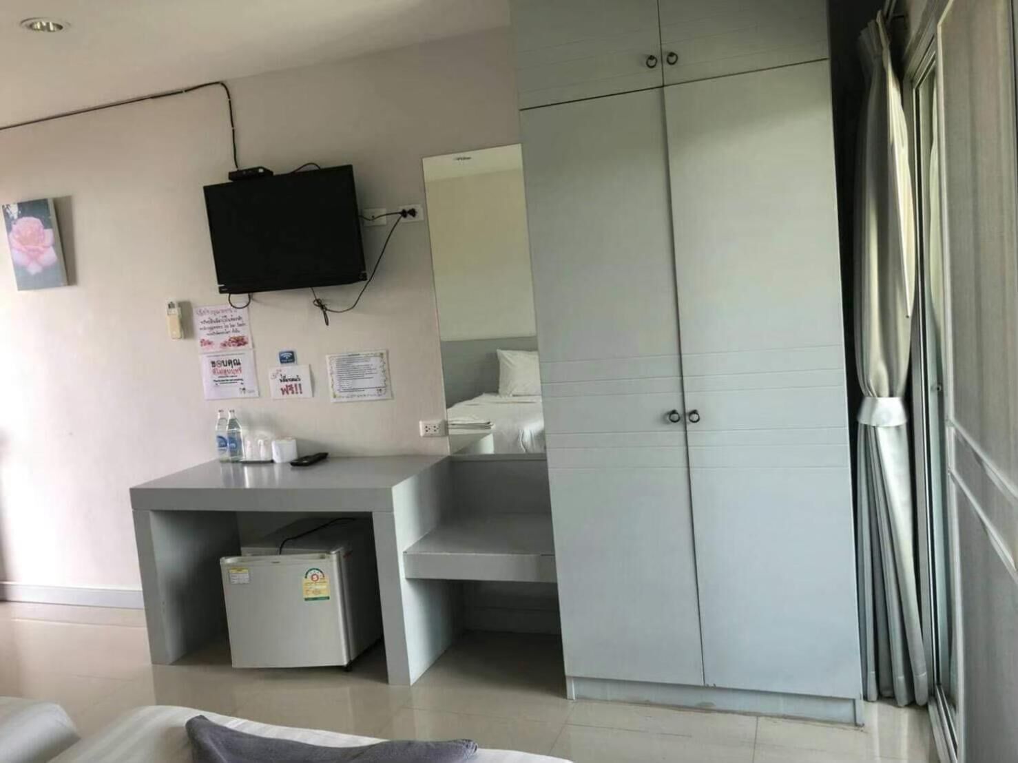 Una camera d'albergo con un armadio grigio, una televisione a schermo piatto fissata al muro, una scrivania con frigorifero e un letto con testiera bianca.