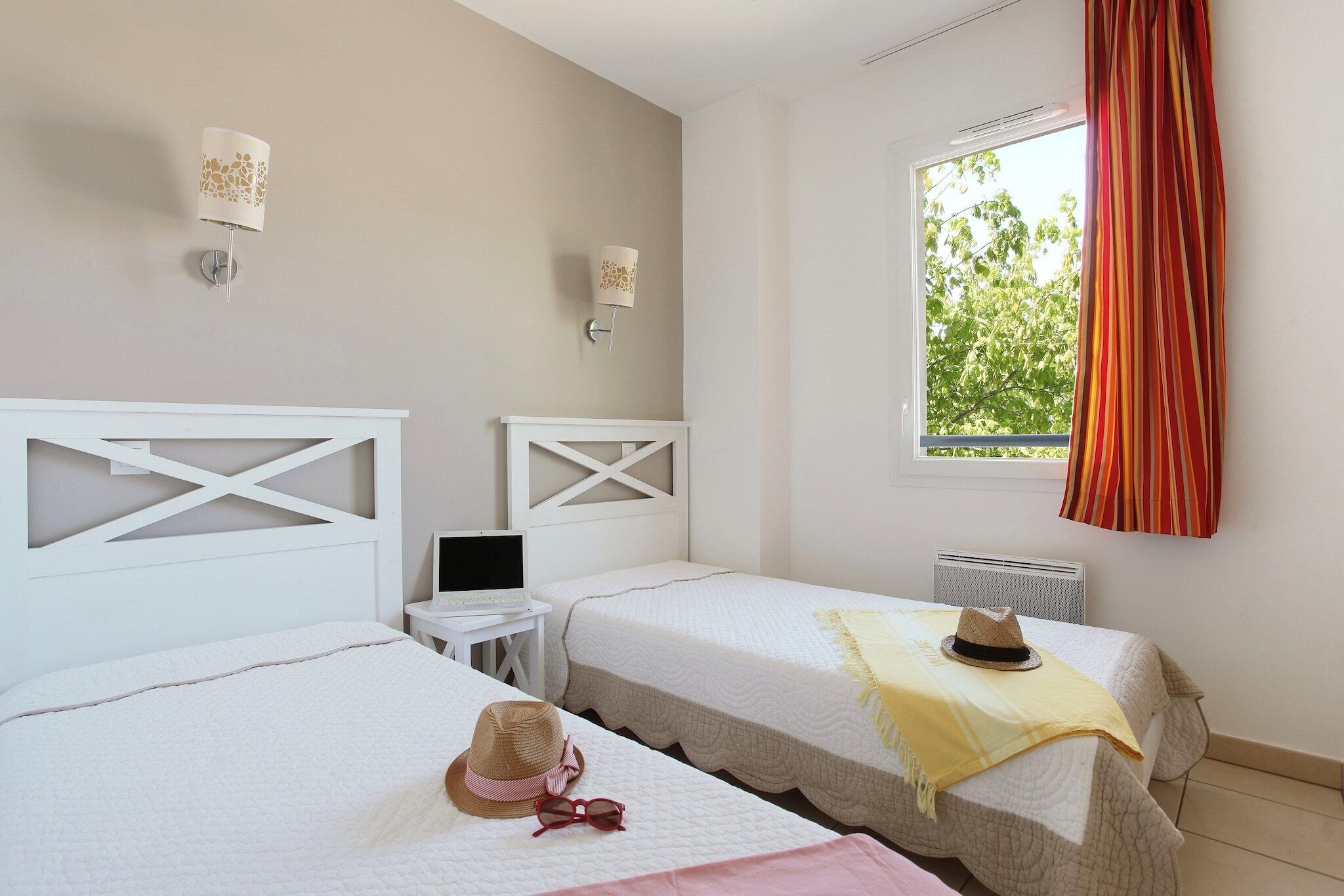 Une chambre d’hôtel avec deux lits, un ordinateur portable posé sur une table d’appoint, une fenêtre avec des rideaux et une vue sur la verdure à l’extérieur.